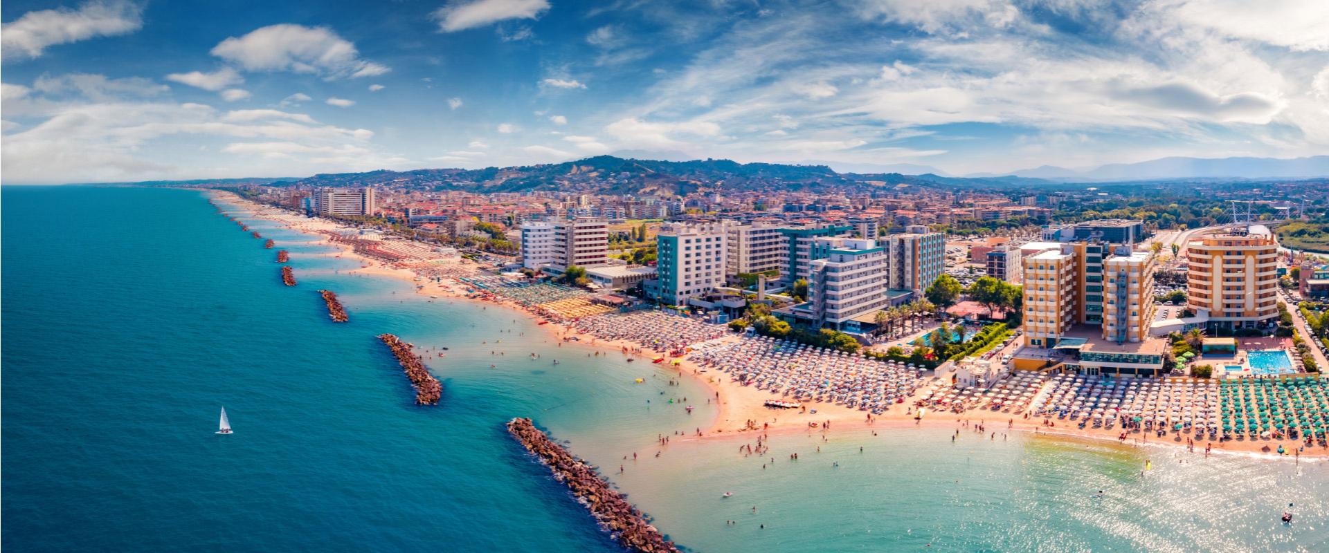 Spiaggia affollata con ombrelloni, edifici moderni e mare calmo in una giornata soleggiata.