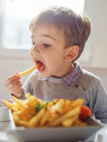Bambino mangia patatine fritte con entusiasmo, seduto a tavola in cucina.